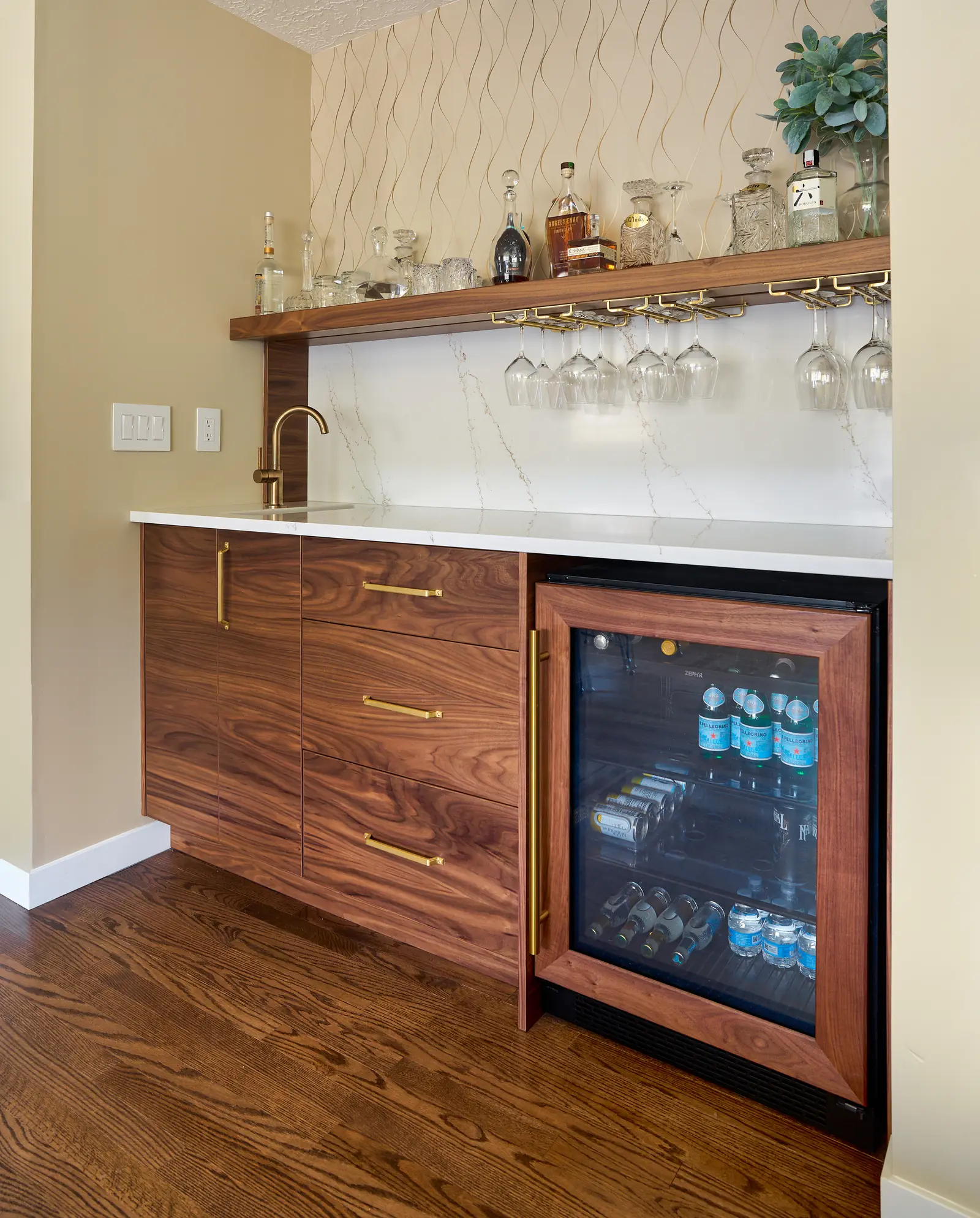 Walnut bar cabinetry with a quartz backsplash, brass stemware rack and a panelled beverage fridge